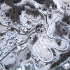 An overhead shot captures the construction of the new Cortina Sliding Centre, a track spanning approximately 1.4 kilometers with 16 curves, designed to host bobsleigh, skeleton, and luge events for the Milano Cortina 2026 Winter Olympics. The project has become a focal point for environmental protests after the felling of approximately 500 ancient larch trees in the Ronco forest to clear the path for the concrete structure