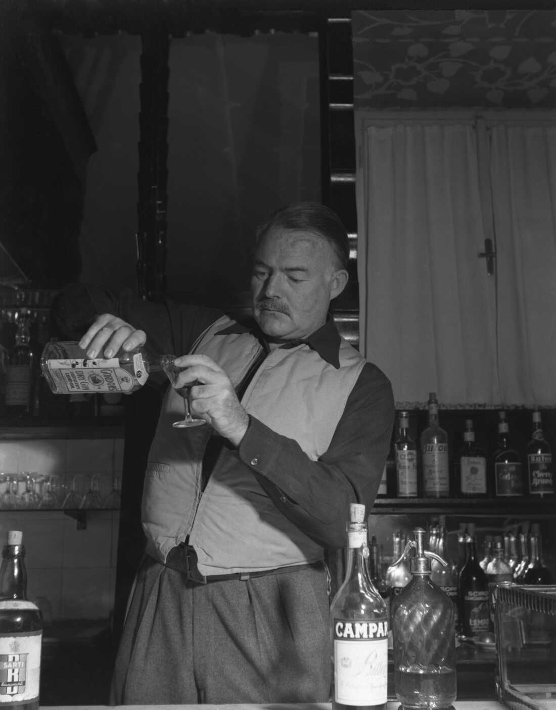 Ernest Hemingway, wearing a hunter waistcoat, stands behind a bar counter and pours gin from a bottle of Gordon's with other bottles of alcohol around him.