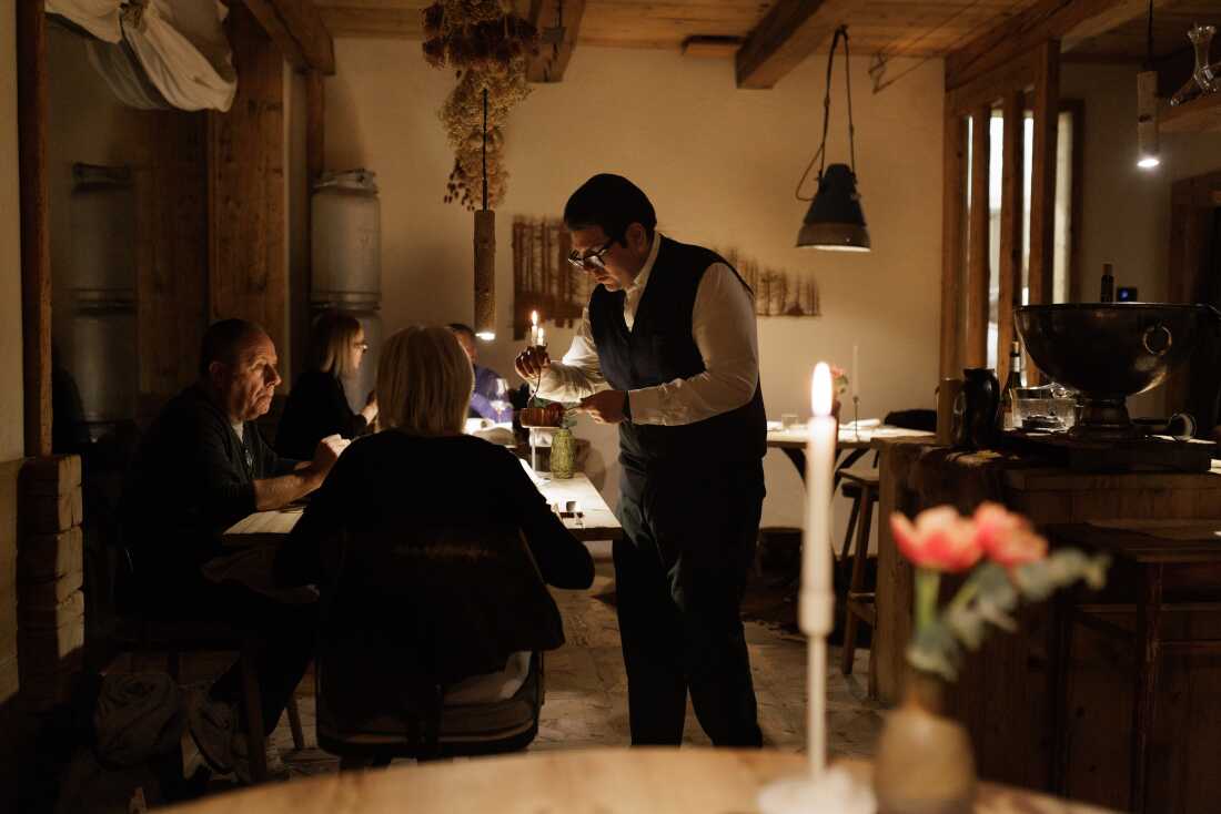 A waiter provides tableside service for guests in the dining room of the Michelin-starred restaurant SanBrite. The establishment, known for its "agricucina" philosophy, combines a refined mountain atmosphere with traditional Cortinese architectural elements, emphasizing a direct connection between local ingredients and high-end hospitality in the heart of the Dolomites.