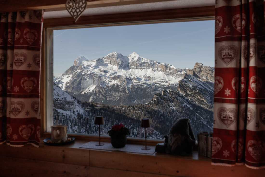 The snow-capped peaks of the Tofane massif are framed through a window of a traditional alpine "Rifugio," decorated with typical heart-patterned Ampezzo textiles. These high-altitude mountain huts serve as essential rest points for skiers and hikers, offering a blend of rustic hospitality and panoramic views that define the winter experience in the UNESCO World Heritage Dolomites.