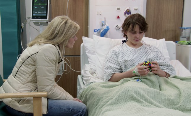 Leanne sitting next to Sam's hospital bed leaning in to talk to him while he stares at a Rubik's Cube in his hands in Corrie