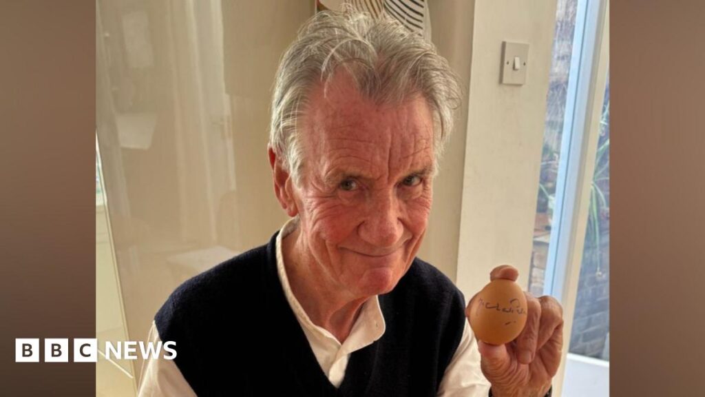 Sir Michael Palin sits at a kitchen table and holds an egg in one hand that he has signed with a pen. He has short grey hair and wears a white shirt with a black vest over the top.