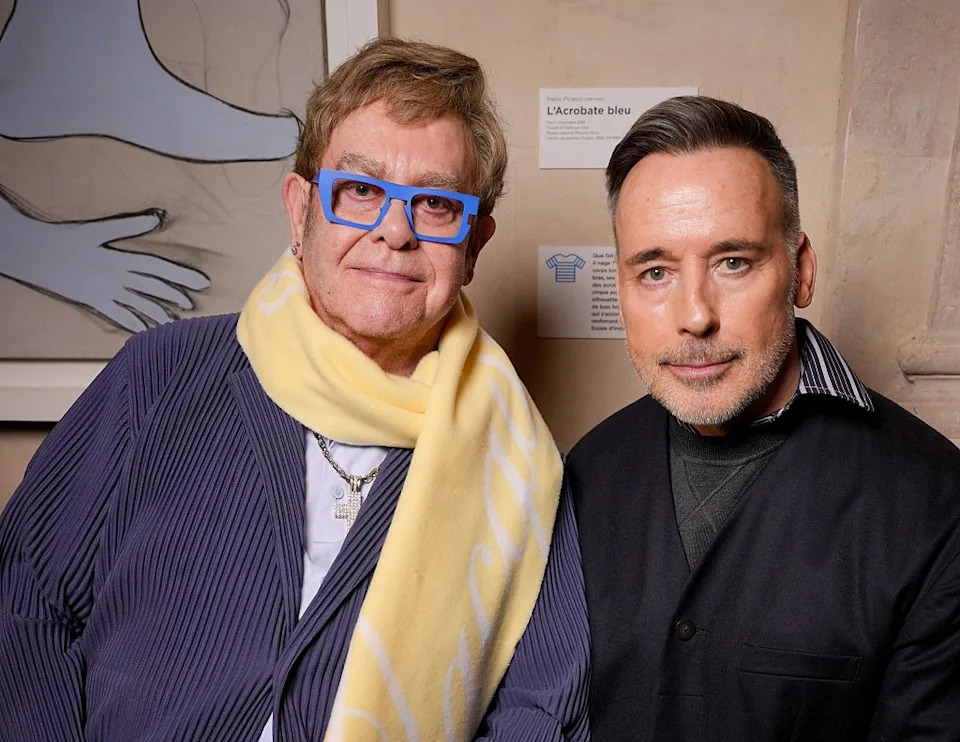 Two individuals posing together; one wears a striped jacket with a scarf, and the other is in a dark jacket. They are in an indoor setting