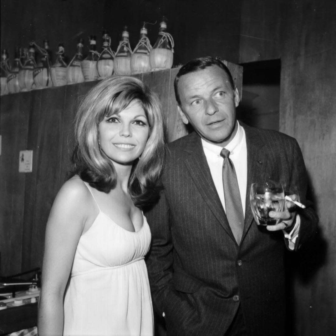 A woman in a white dress stands next to a man in a pinstripe suit holding a glass at a social event, with decorative bottles displayed on a high shelf behind them.