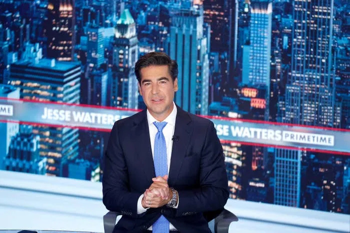 A person in a suit sits at a news desk, cityscape backdrop