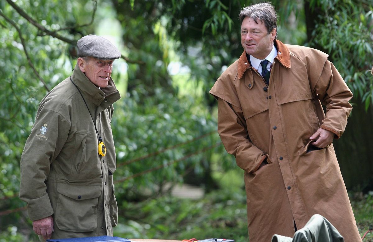 Two men dressed in winter coats are standing in an outdoor setting surrounded by greenery. The individual on the left wears a cap and a jacket with a yellow emblem on the left chest, while the person on the right dons a long coat and a tie. The background features lush trees and plants, indicating a park or garden environment.