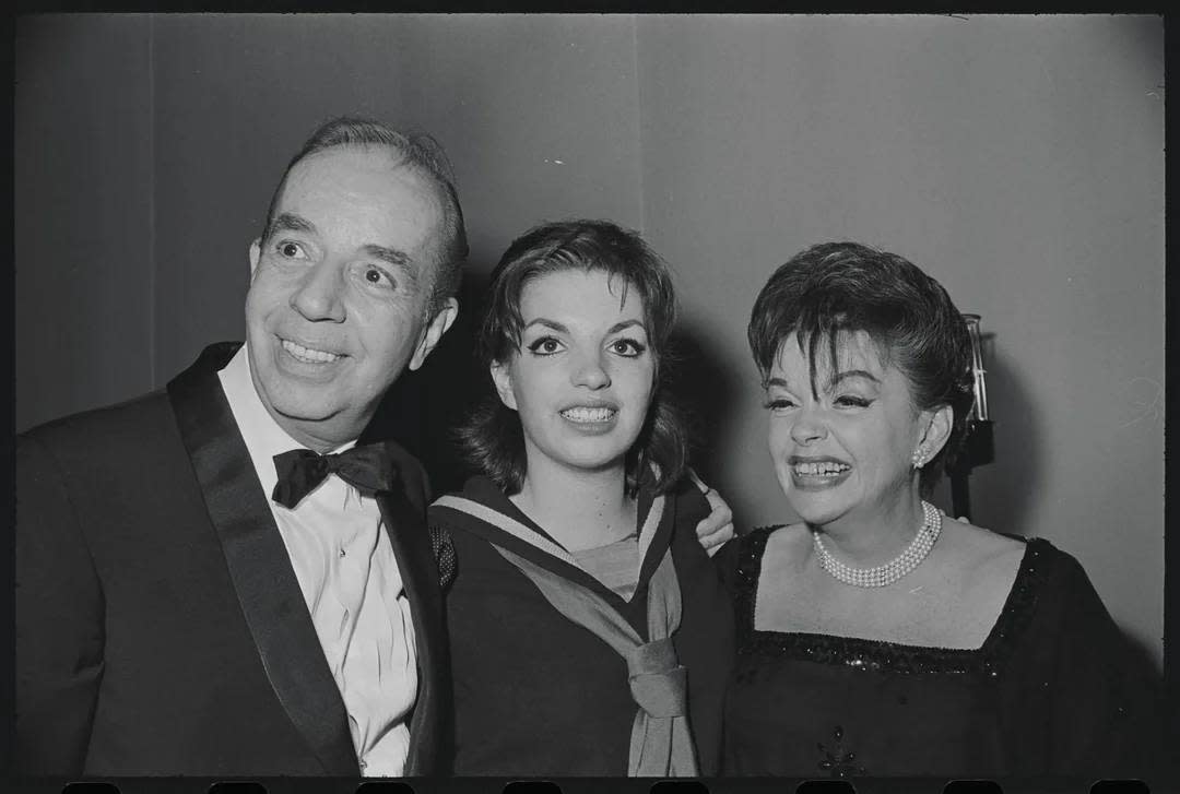 Three people dressed in formal attire stand together, smiling for the camera at what appears to be an event or gathering. The background is plain, and all three are looking forward.