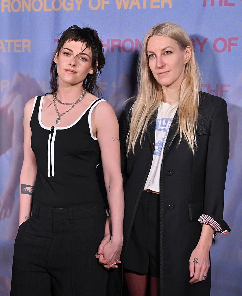 Two individuals holding hands at an event, one wearing a sleeveless top with necklaces and the other in a coat over a graphic tee