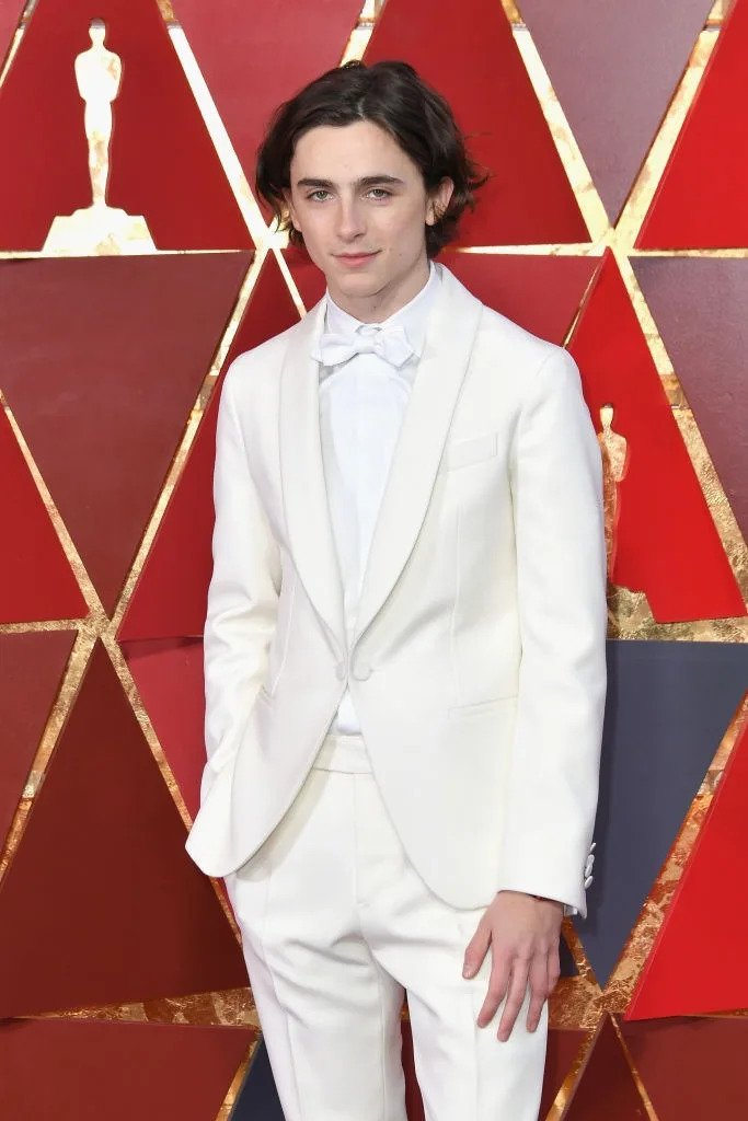 Person in a white tuxedo with a bow tie on a red carpet with geometric backdrop