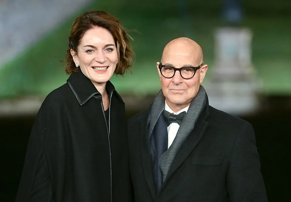 A couple smiles while dressed in elegant, formal attire, standing outdoors