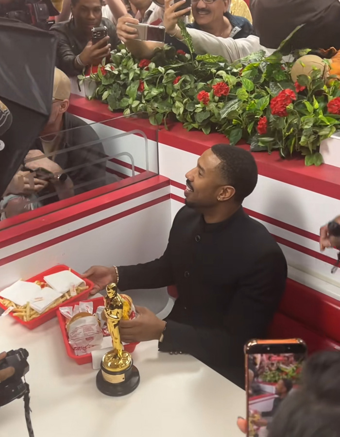 Michael B. Jordan smiling at a fast food restaurant holding an Oscar statue while fans take photos nearby.