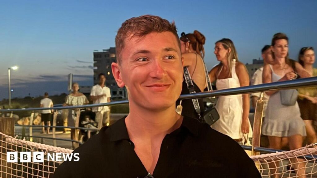 ‘Everything has changed’: Missile attacks shatter Dubai’s safe haven image Will Bailey looks to the side as he smiles. He is sitting down wearing a black shirt. People can be seen walking on a walkway behind him.