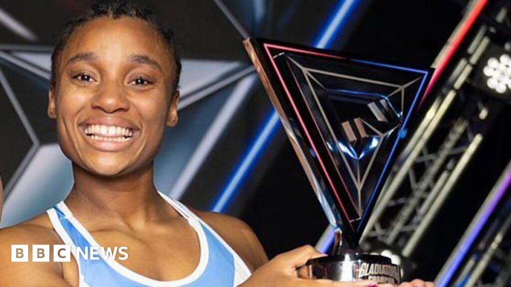 Gladiators 2026: Croydon’s Emily Bell crown champion Close-up of Emily Bell smiling and holding up her trophy. She is wearing a blue and white tank top and has plaited dark hair.