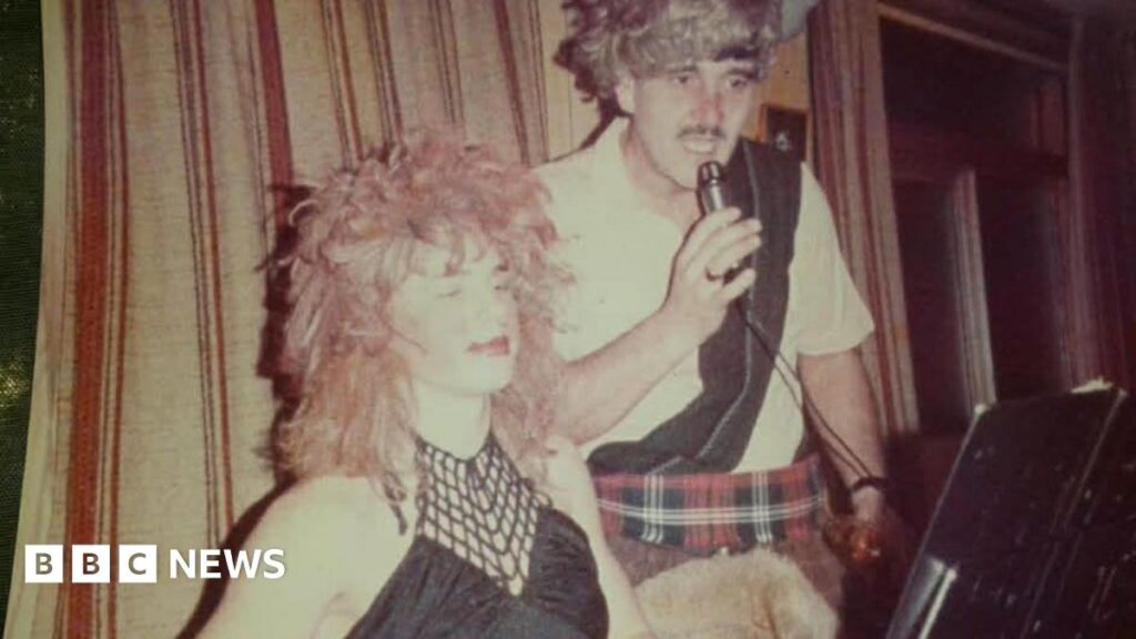 Take That’s Gary Barlow’s left clubgoers ‘gobsmacked’ at 11 years old Barlow and Trevor Davies in fancy dress on stage. Barlow wears a long red wig and black woman's top, while Davies wears red tartan kilt, white shirt with a black sash, and a grey wig. Davies holds a microphone, with a stage curtain behind them both.