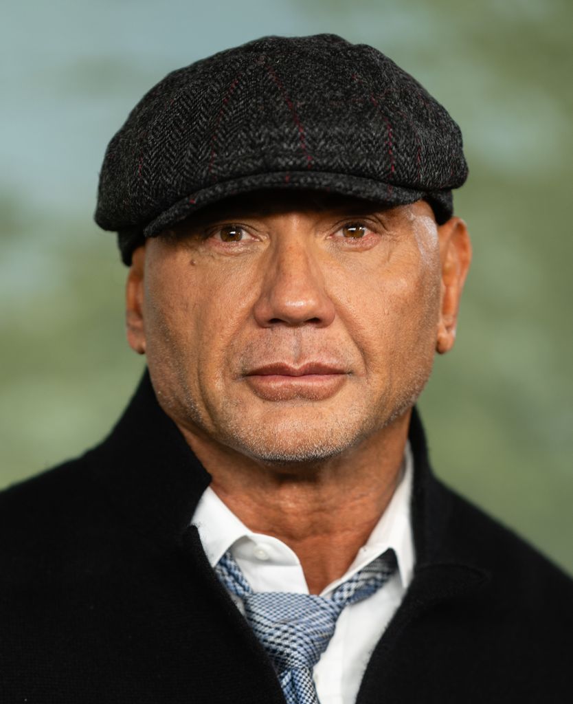 Dave Bautista wears a shirt, black jacket and flat cap and looks straight down the camera lens.