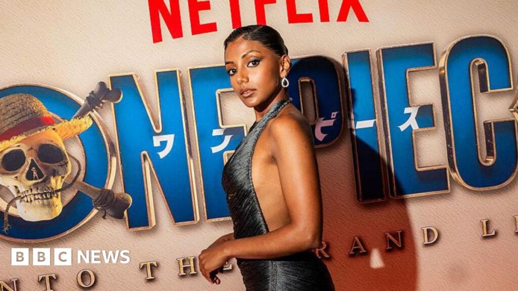 Charithra Chandran wears a metallic strapless dress and has black hair tied up in a bun, with a silver earring. She stands in front of a sign on the red carpet for the One Piece premiere