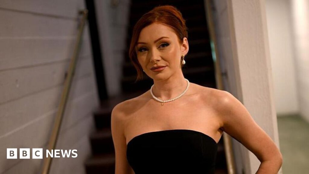 Elz is looking straight ahead at the camera standing at the bottom of some stairs. She has her red hair tied up and is wearing a black strapless dress. She has a pearl necklace on and pearl earrings.