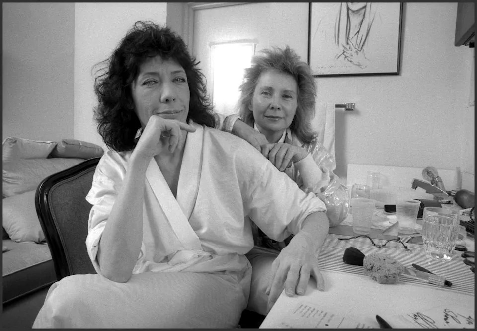Two women in a casual, intimate setting; one is seated in a robe with an arm resting on a table, while the other stands behind, both appearing relaxed
