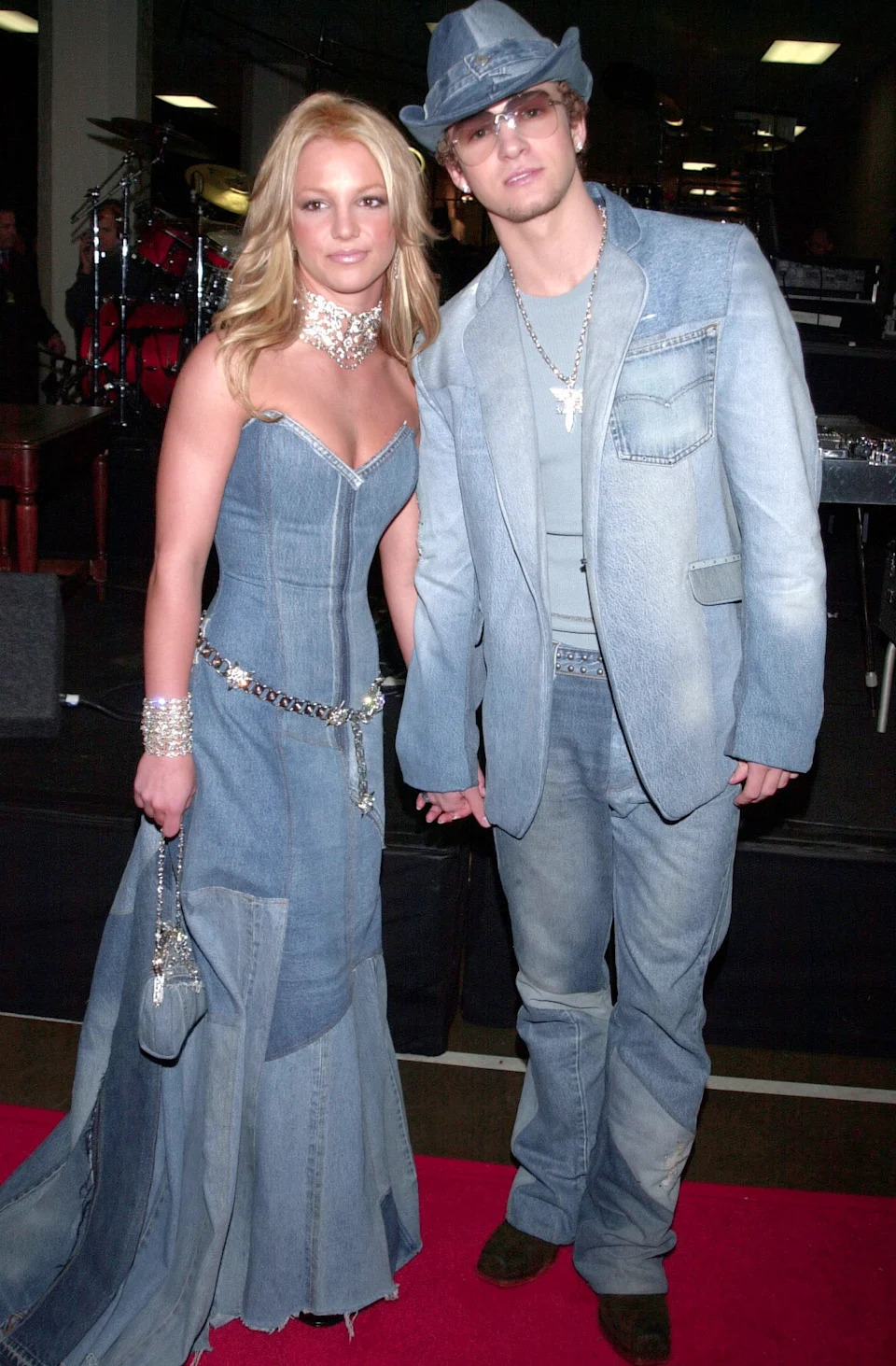 Two individuals on a red carpet wearing matching denim outfits, including dresses and hats, accessorized with jewelry