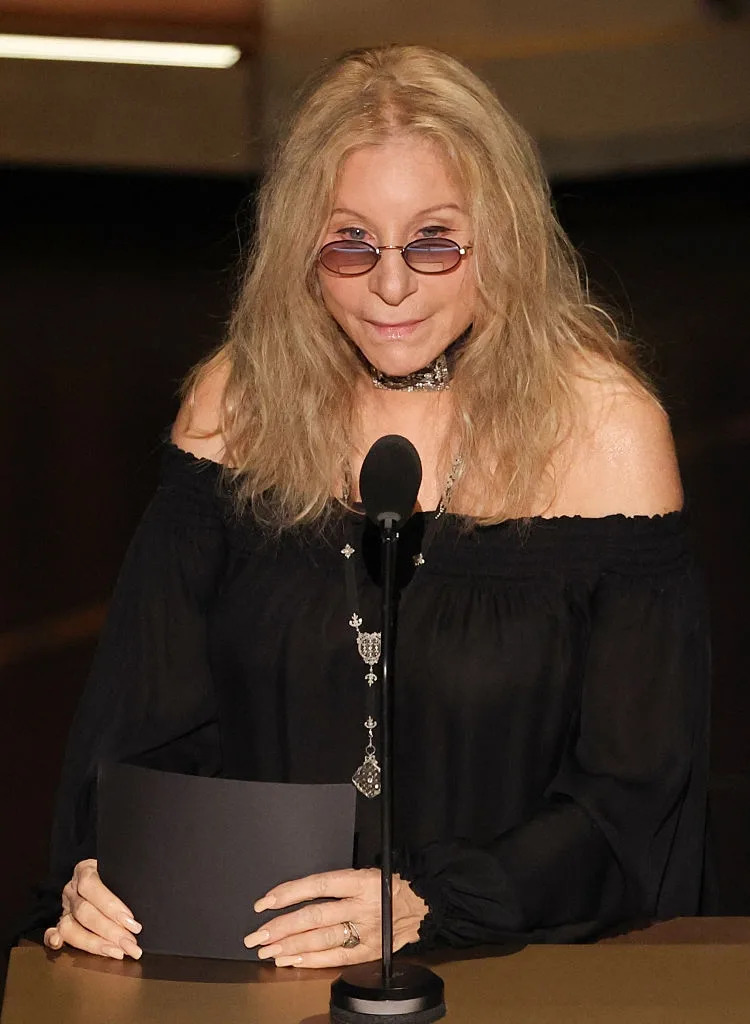 Person in an off-the-shoulder dress, wearing sunglasses and a necklace, stands at a podium, holding a card during a formal event