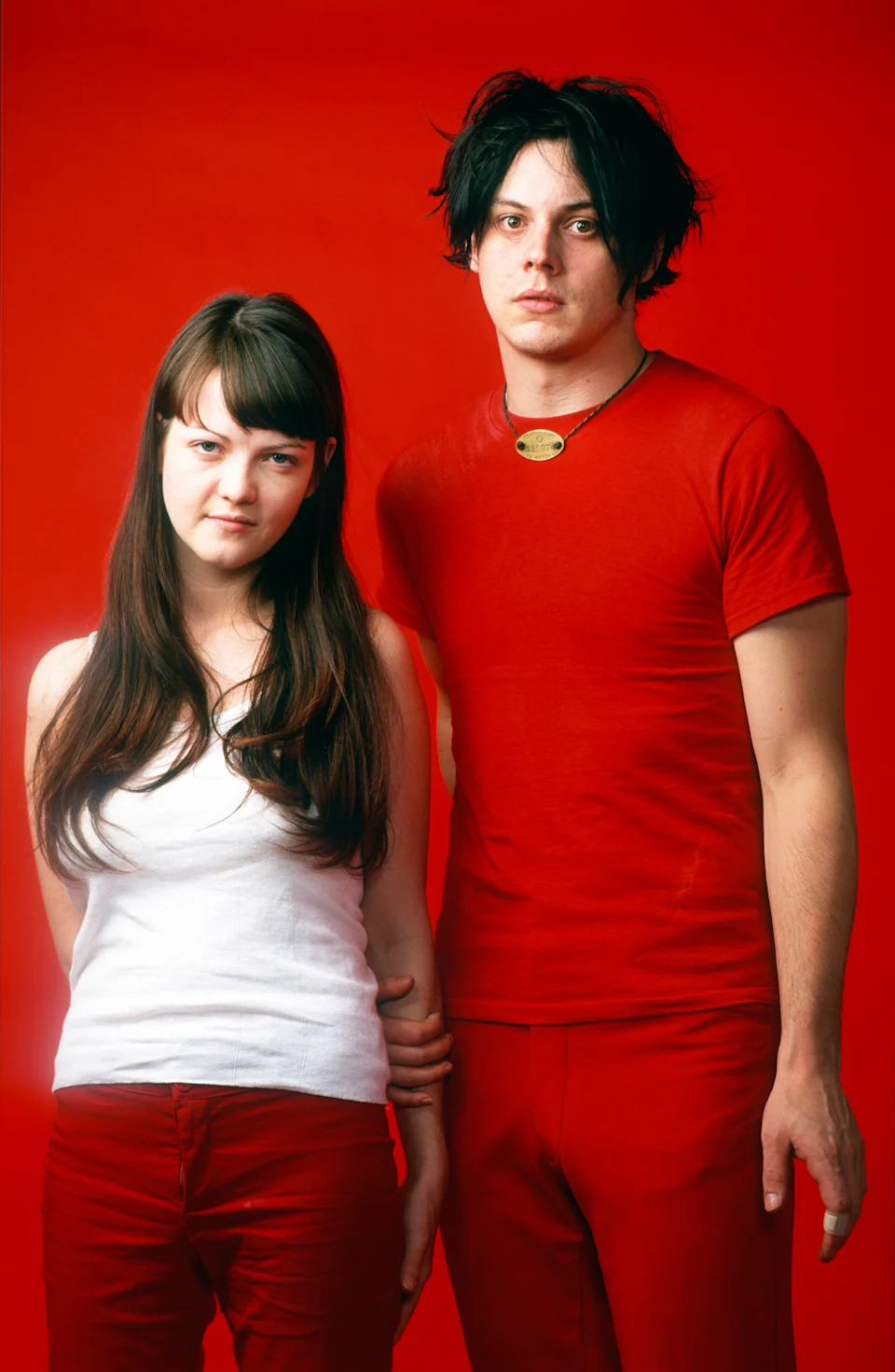 Two people pose in front of a plain backdrop, one in a white top and red pants, the other in a red shirt and pants
