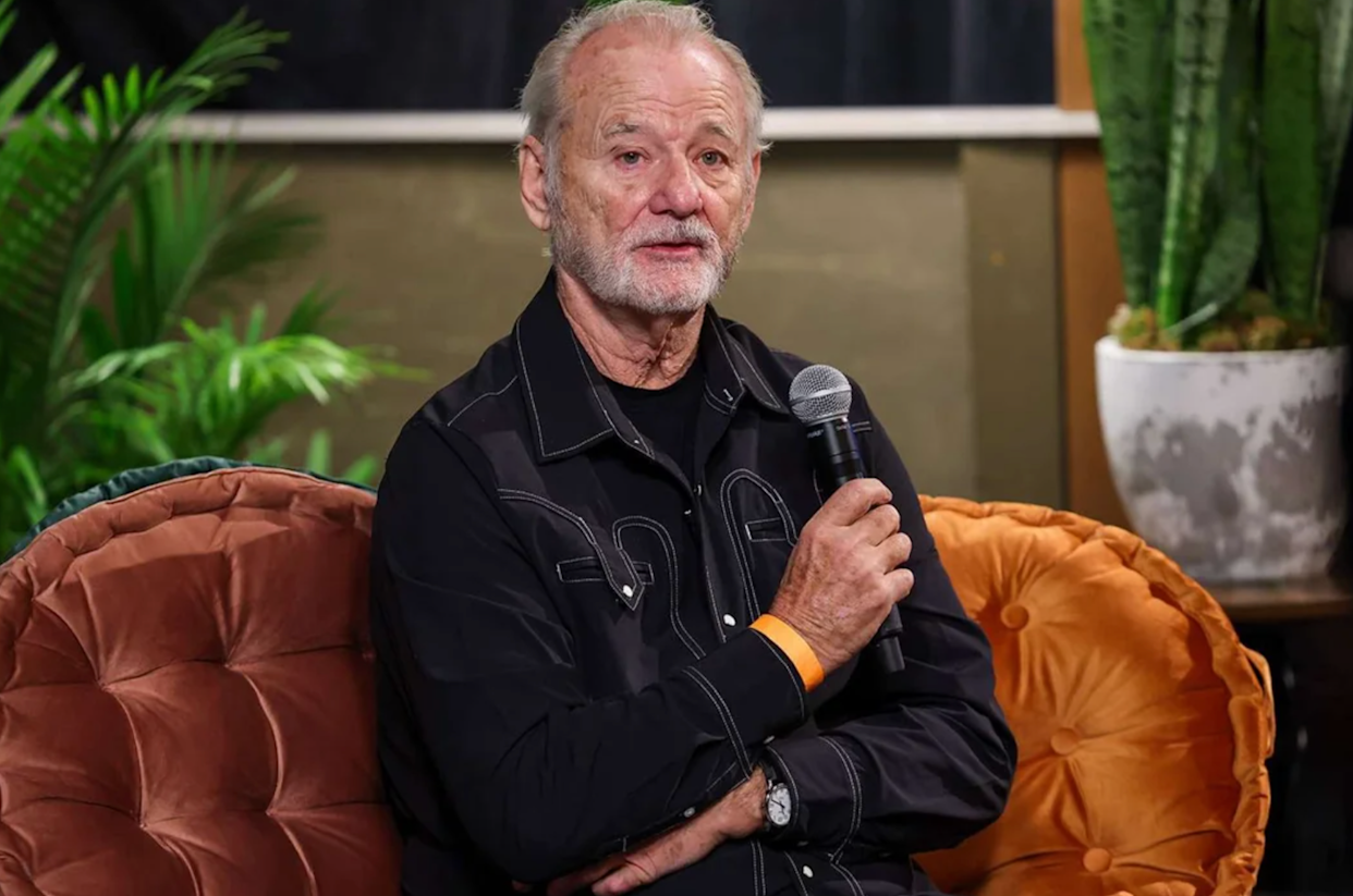 An older man with gray hair and a beard sits on a brown couch, holding a microphone in his right hand. He is wearing a black shirt with white stitching. Green plants are visible in the background.