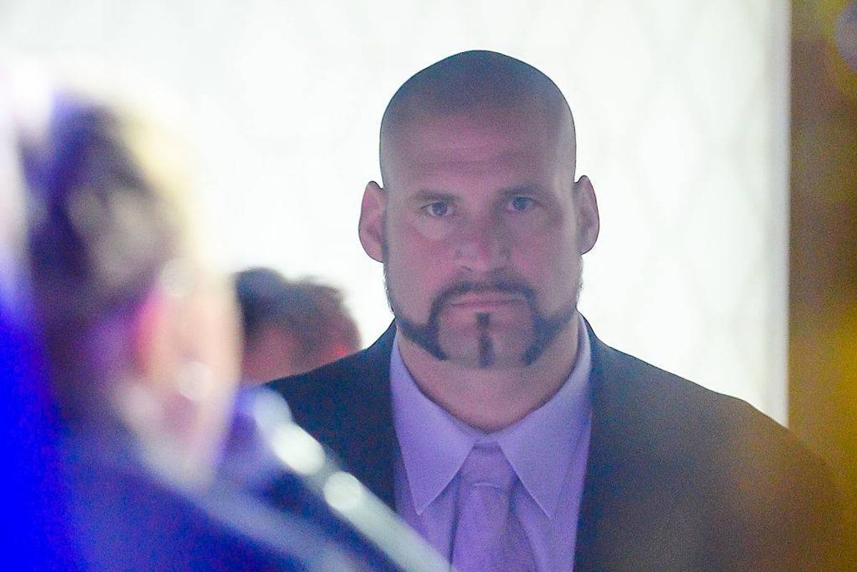 An individual dressed in formal attire, including a suit and tie, stands in a well-lit indoor setting, facing another person. The person's facial features, including a mustache and beard, are prominently visible, with a blurred figure in the background.
