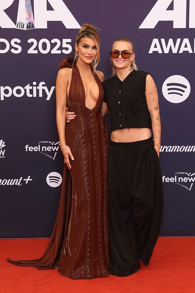 Two individuals posing on a red carpet. One in a plunging gown, the other in a sleeveless top and pants with tattoos