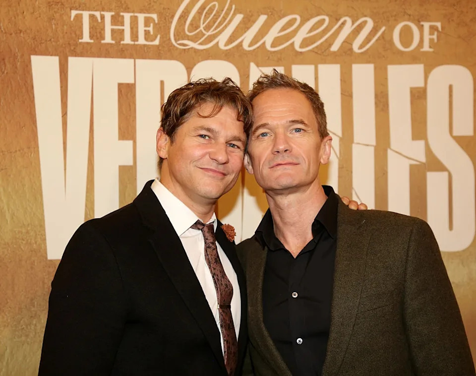 Two people in formal attire stand closely, posing in front of "The Queen of Versailles" backdrop at a public event