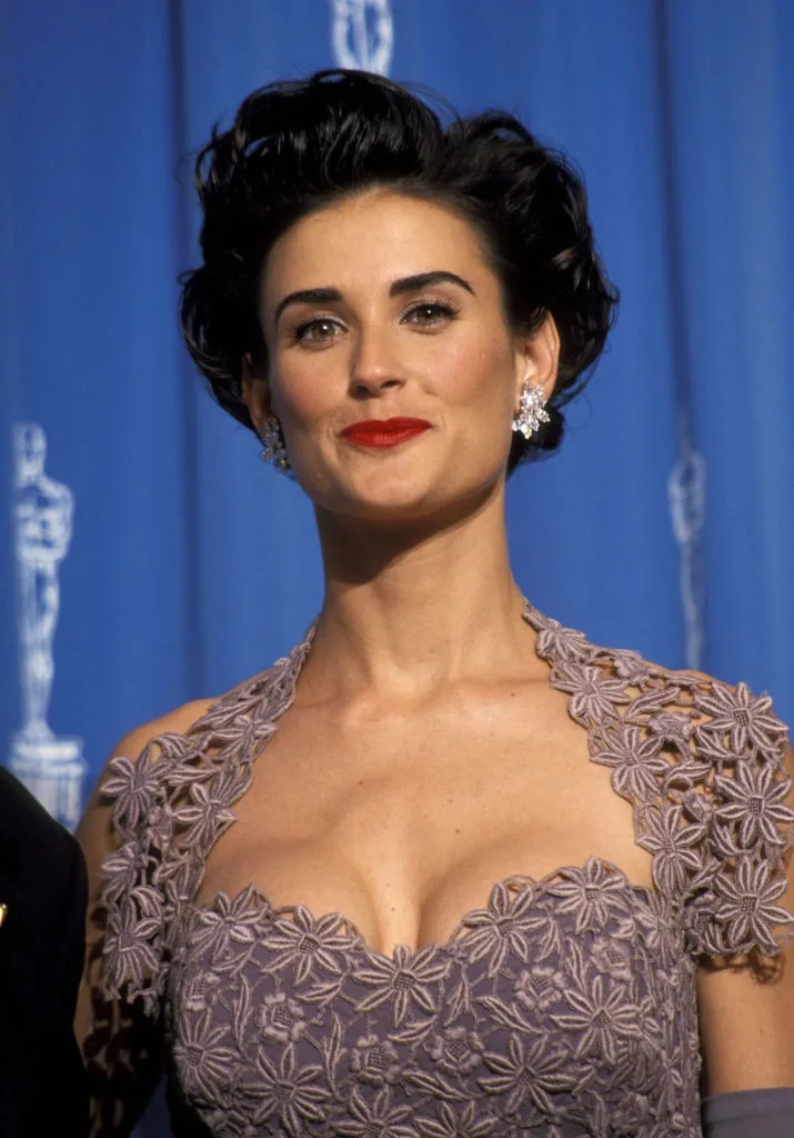 Woman with short styled hair in an elegant floral lace dress, attending a formal event with a backdrop featuring an Oscar statue