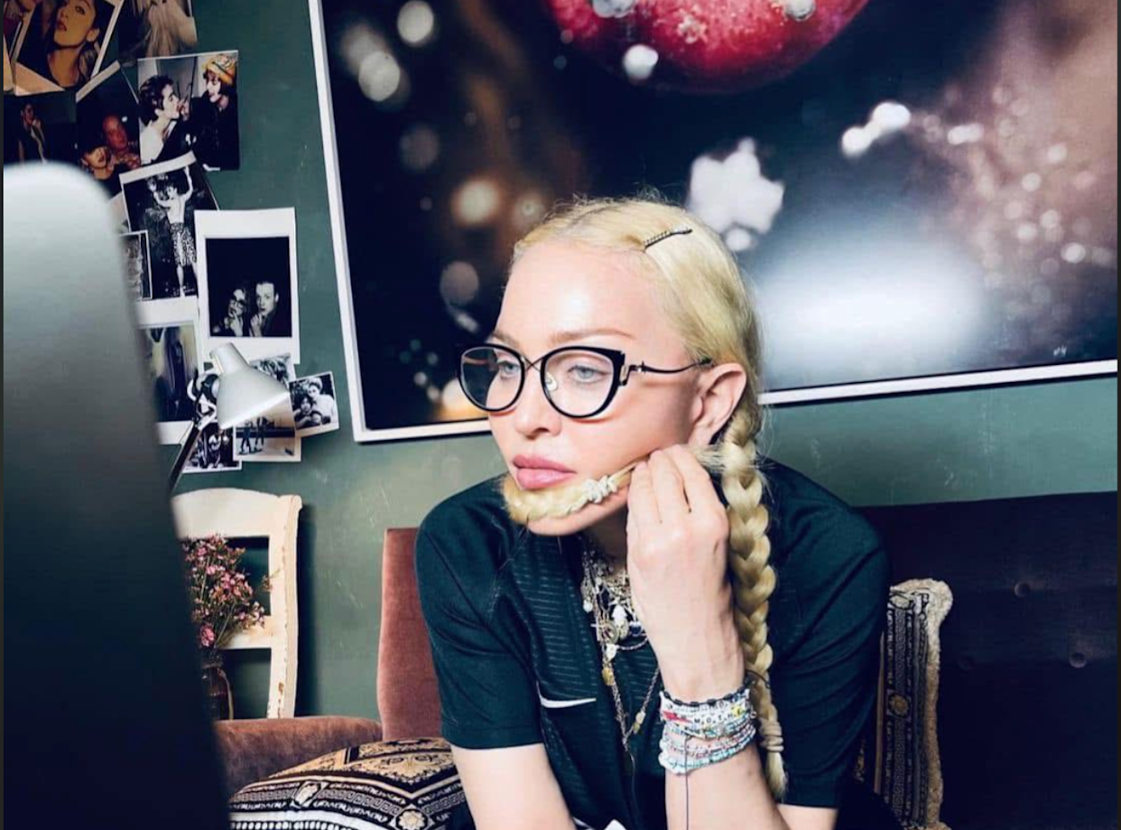 A woman with long blonde braids and glasses sits indoors, resting her chin on her hand and looking at a laptop. She has several bracelets on her wrist. Behind her are photos on the wall and a large abstract print.