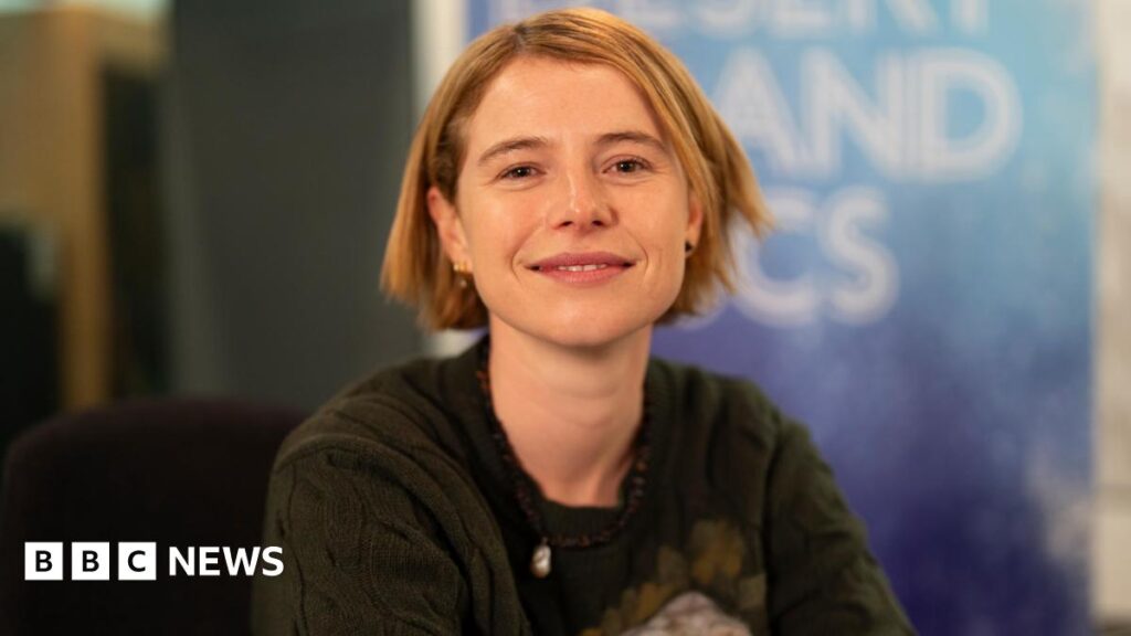 Jessie Buckley, who has short blonde hair and pierced ears with different earrings in. She is wearing a dark green knitted jumper and a bead necklace. She is smiling at the camera and sitting in front of a Desert Island Discs backdrop