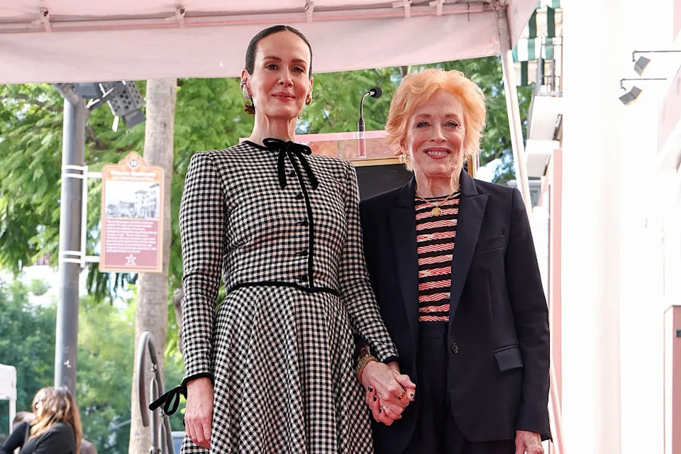 Two individuals pose together outdoors, holding hands. One wears a checkered dress, the other is in a striped top and blazer
