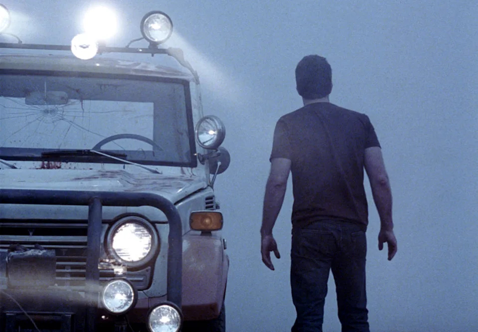 A man stands facing a classic SUV with headlights piercing through fog, creating a mysterious atmosphere