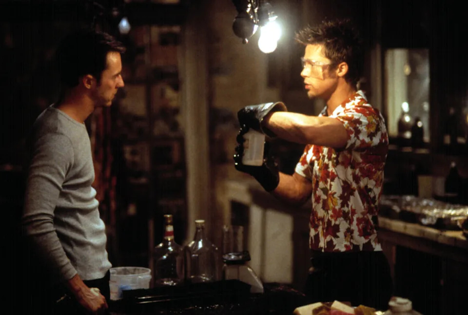 Two men in a dimly lit room; one wears a floral shirt and rubber gloves, appearing to instruct the other. They are surrounded by bottles and jars