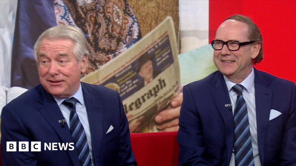 Entertainers Ian Ashpitel (left) and Jonty Stephen (right) on the BBC North West Tonight studio sofa. They are both wearing matching navy blue suits with matching ties. There is a Telegraph newspaper in the background which is part of a sketch from their show.