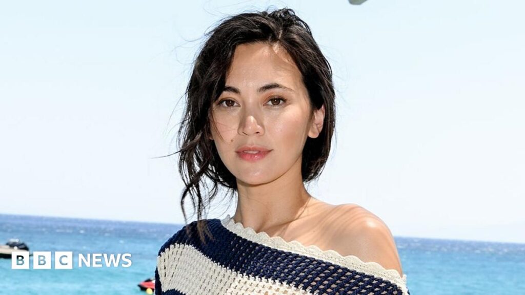 A still from How to Make a Killing shows Jessica Henwick, with her hair tied up and paired with a strappy party dress, giving the side-eye to an individual off-screen. She's bathed in a moody, candle-esque light.