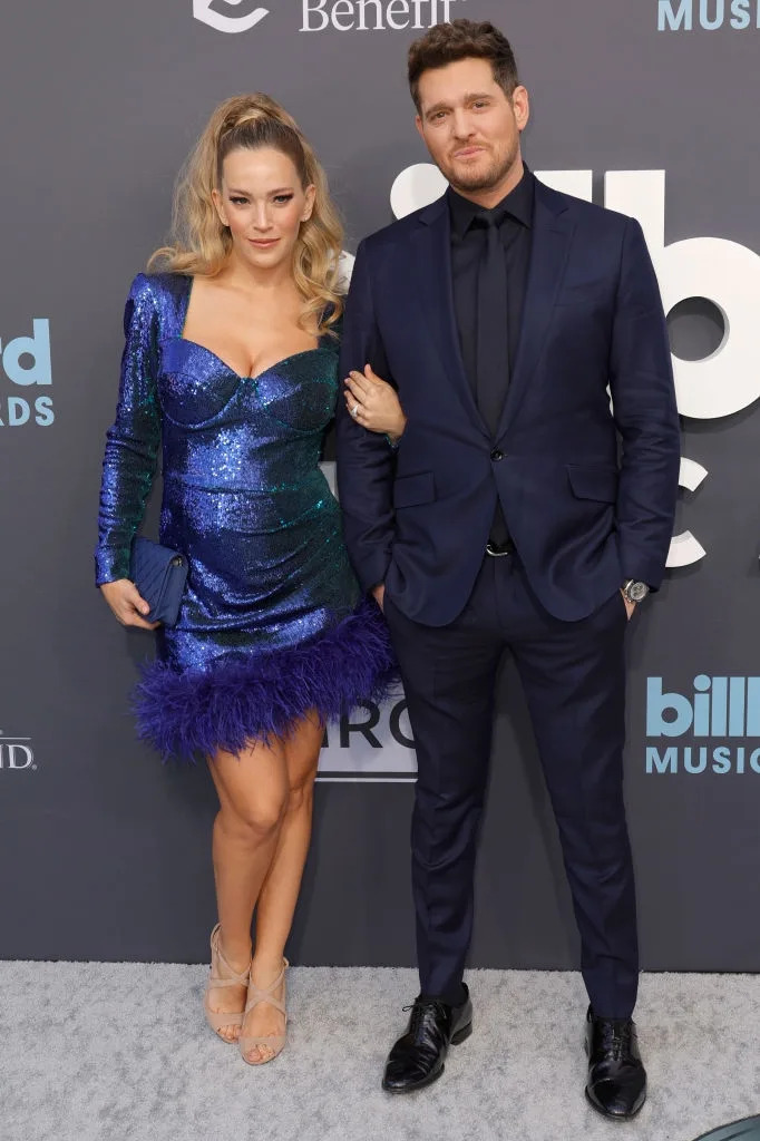 A couple poses on the red carpet; the woman wears a sparkling dress with feather trim, and the man is in a classic suit and tie