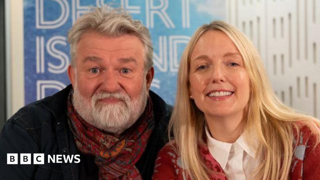 Si King and Lauren Laverne smiling into the camera in front of a blue Desert Island Discs sign. Si is smiling with his lips pursed and has white, short hair and a white beard. He is wearing a black jacket and red paisley scarf which is tied in a knot around his neck. Lauren Laverne is sitting to the right of him (as you look towards them) and has long, blonde hair. She is smiling with her teeth and has glossy lips. She is wearing a red and blue patterned cardigan with a white shirt underneath with is buttoned all the way to the top.