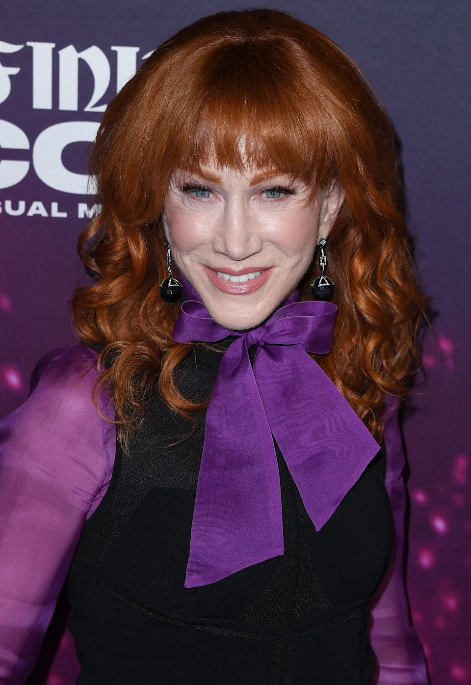 Person with red curly hair and bangs, wearing a black outfit with a large purple bow and dangling earrings, smiling at an event