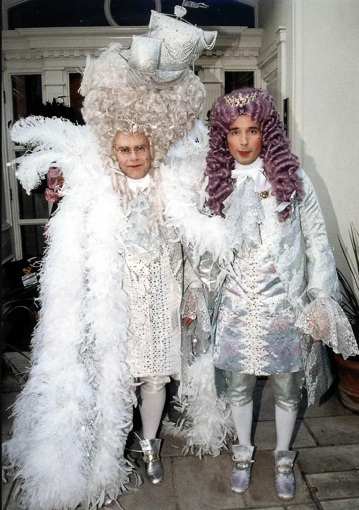 Two individuals in elaborate, historical costume attire with ornate wigs and embellished outfits, standing together at a social event
