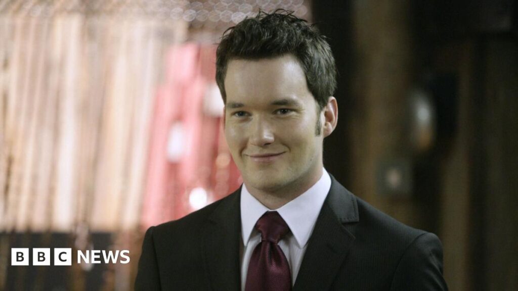 A wall covered in pictures and a string of Welsh flags. At the top a board reads Ianto's Shrine in white writing. The pictures are attached to a rusty metal grid.