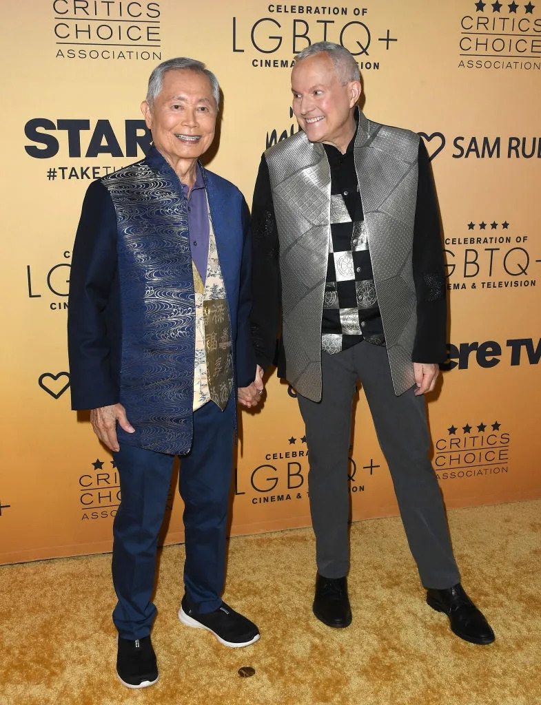 Two people smiling and holding hands on a red carpet. Both wear stylish jackets with unique patterns. Event backdrop reads "Critics Choice Association."