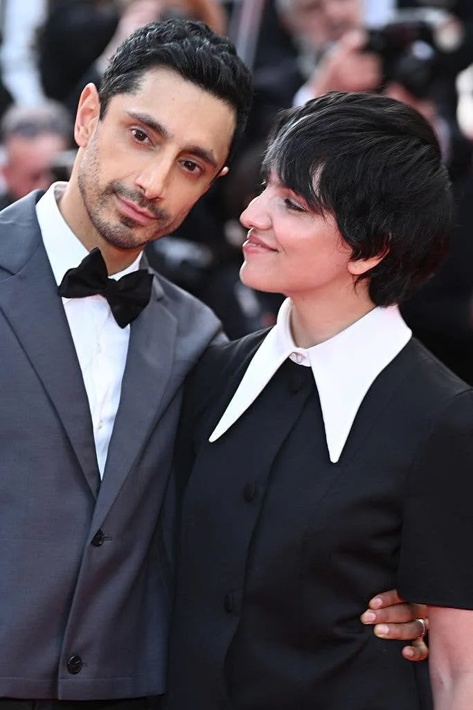 Two individuals on a red carpet. One wears a sleek tuxedo; the other is in a dress with a large white collar, looking at the other person affectionately