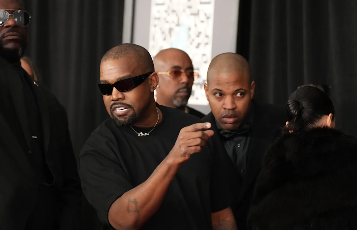 A man in a black t-shirt and sunglasses gestures while standing among several other people, all dressed in dark clothing, at an indoor event with a black backdrop.