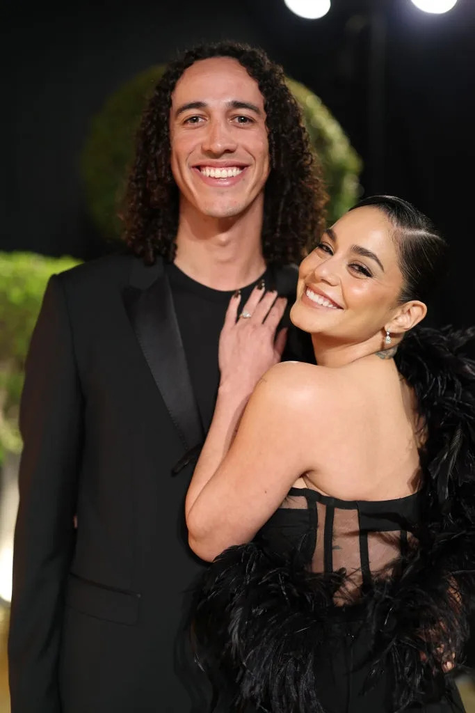 Two people smiling, one in a sleek black suit and the other in a black dress with feather details, posing closely on a red carpet
