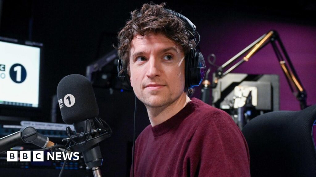 Greg James shares challenge update after his dad suffers stroke Greg James is wearing a burgundy jumper and is wearing headphones on his head. He is sitting at a desk in front of a microphone with a Radio 1 muff.