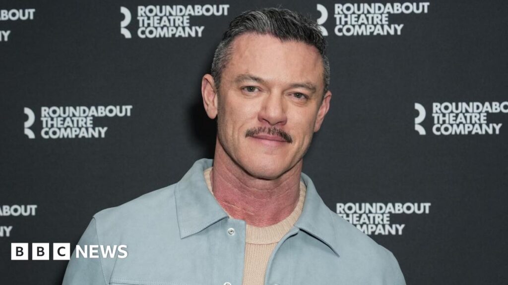 Headshot of Luke Evans. He wears a light blue jacket and cream jumper.  He has greying hair and a moustache