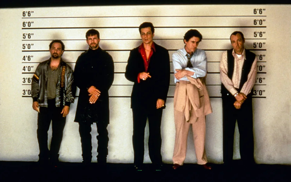 Five men stand against a police lineup wall, dressed in casual to formal attire, resembling a scene commonly related to crime movies