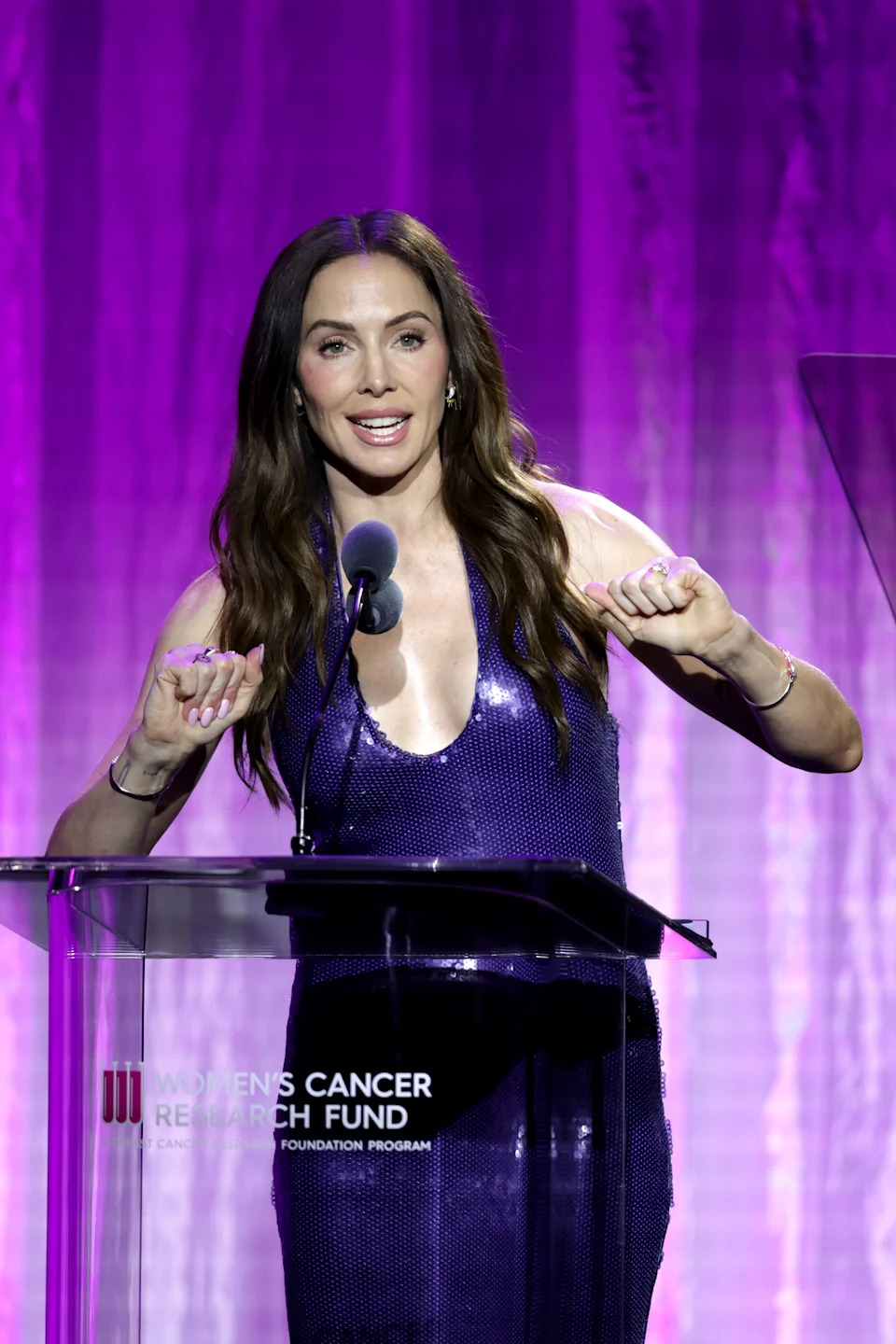 Whitney Cummings speaking at a podium with a microphone, wearing a purple sequined gown, with 'Women’s Cancer Research Fund' text on the lectern and a purple backdrop.